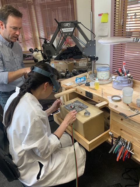 woman in lab coat sitting at wooden work station holding soldering iron
